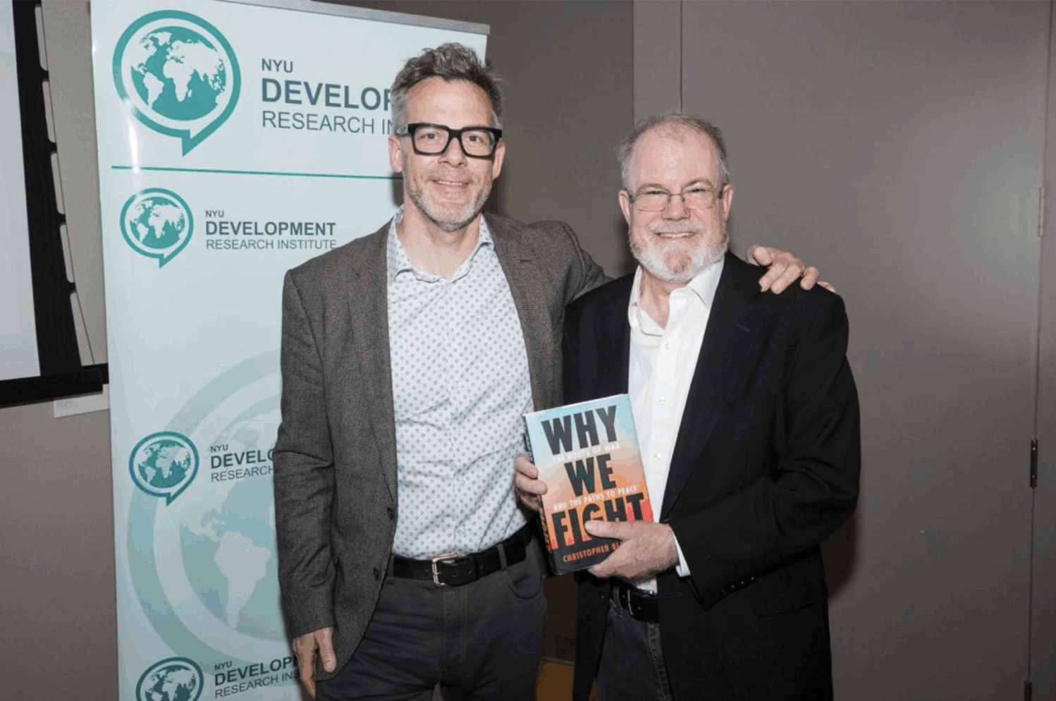 Two men stand and smile at the camera; one holds a book titled Why We Fight. Behind them is a banner with the NYU Development Research Institute logo. A laptop screen with various application tabs is visible.