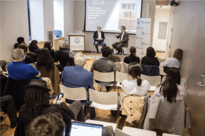 A group of people sit facing two speakers at the front of a room during a presentation. The projected screen displays the words “WHY WE FIGHT.” Laptops and notebooks are visible.