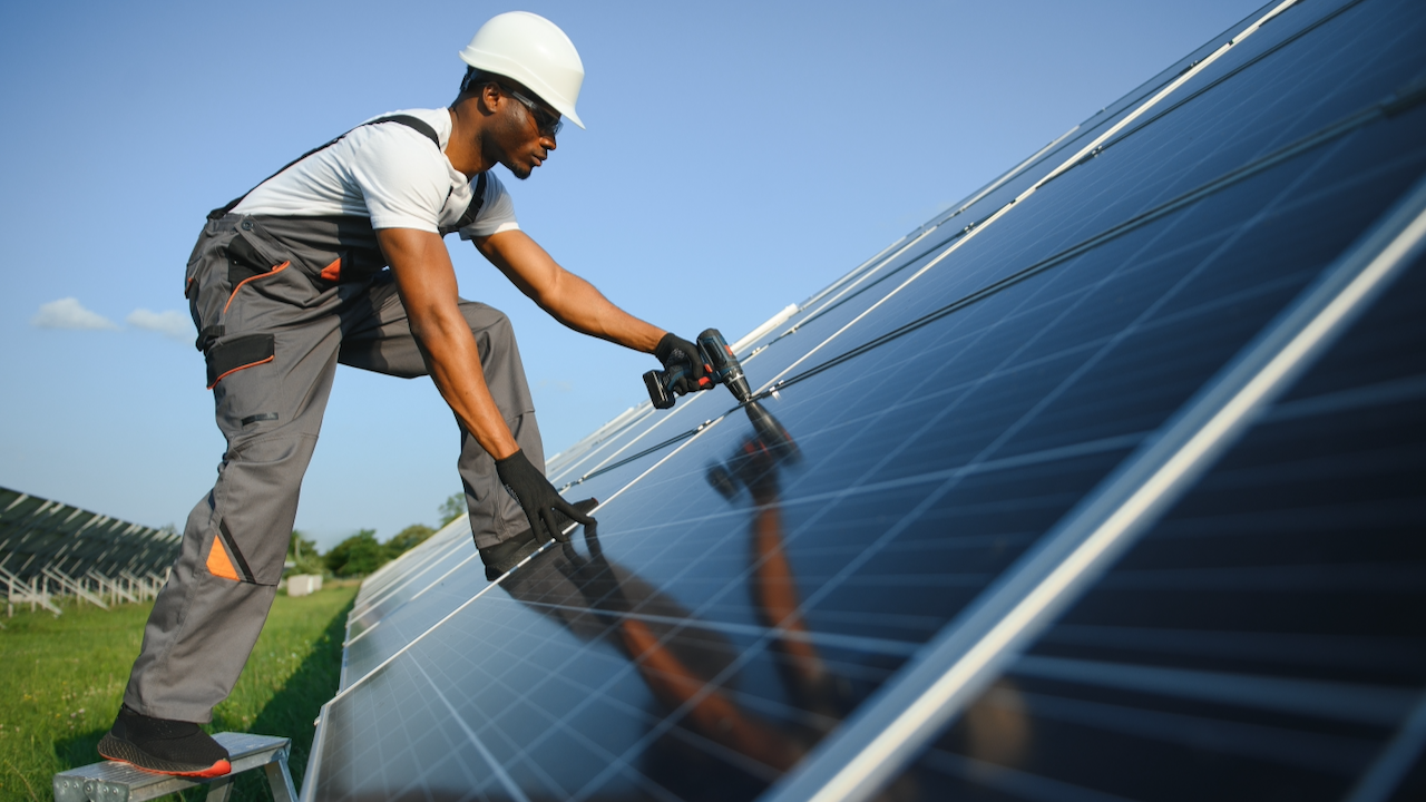 A worker in safety gear installs or maintains a solar panel outdoors on a sunny day, using a tool while standing on a ladder next to a large array of solar panels.