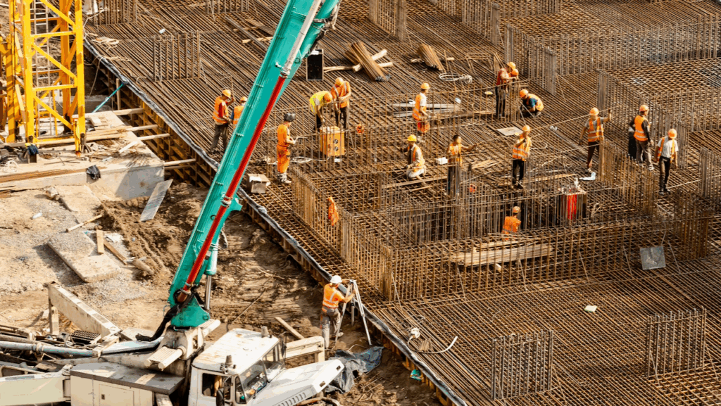 Construction workers in orange vests and helmets work on a large building site, standing on a framework of steel rebar with a crane and concrete pump visible in the muddy area below.