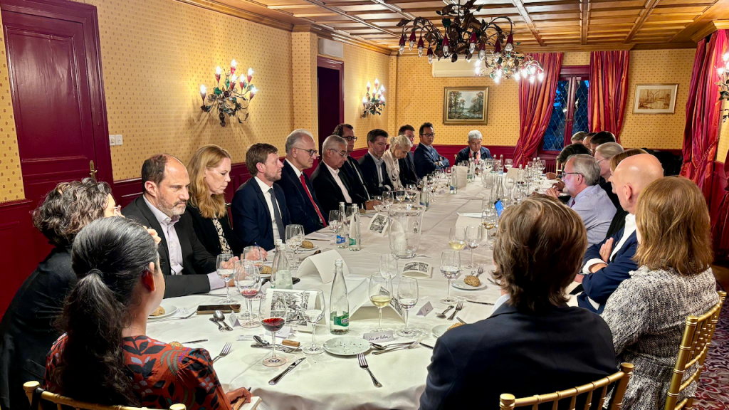 A group of people in formal attire sit around a long, elegantly set dining table in a warmly lit, classic room with red accents, chandeliers, and patterned wallpaper, engaged in conversation.