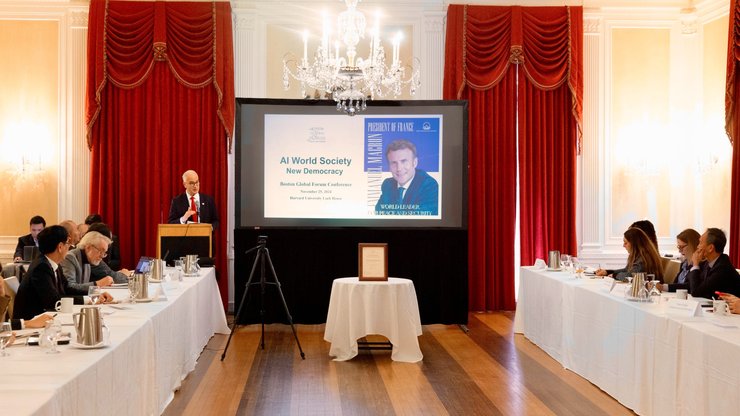 A formal conference room with red curtains and chandeliers; a man speaks at a podium to seated attendees. A large screen displays a slide titled “AI World Society New Democracy” with a photo of a man on the right.