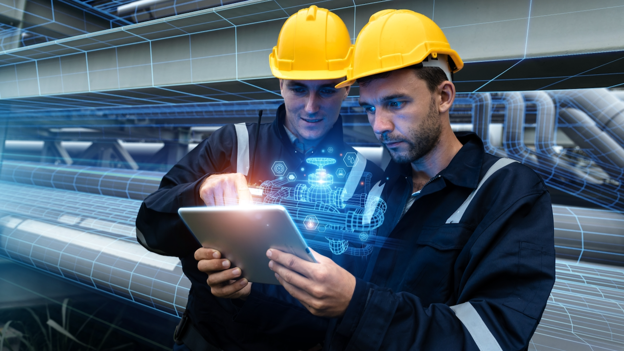 Two engineers in hard hats and work uniforms use a tablet, with digital graphics of machinery overlayed, suggesting they are interacting with virtual or augmented reality in an industrial or factory setting.