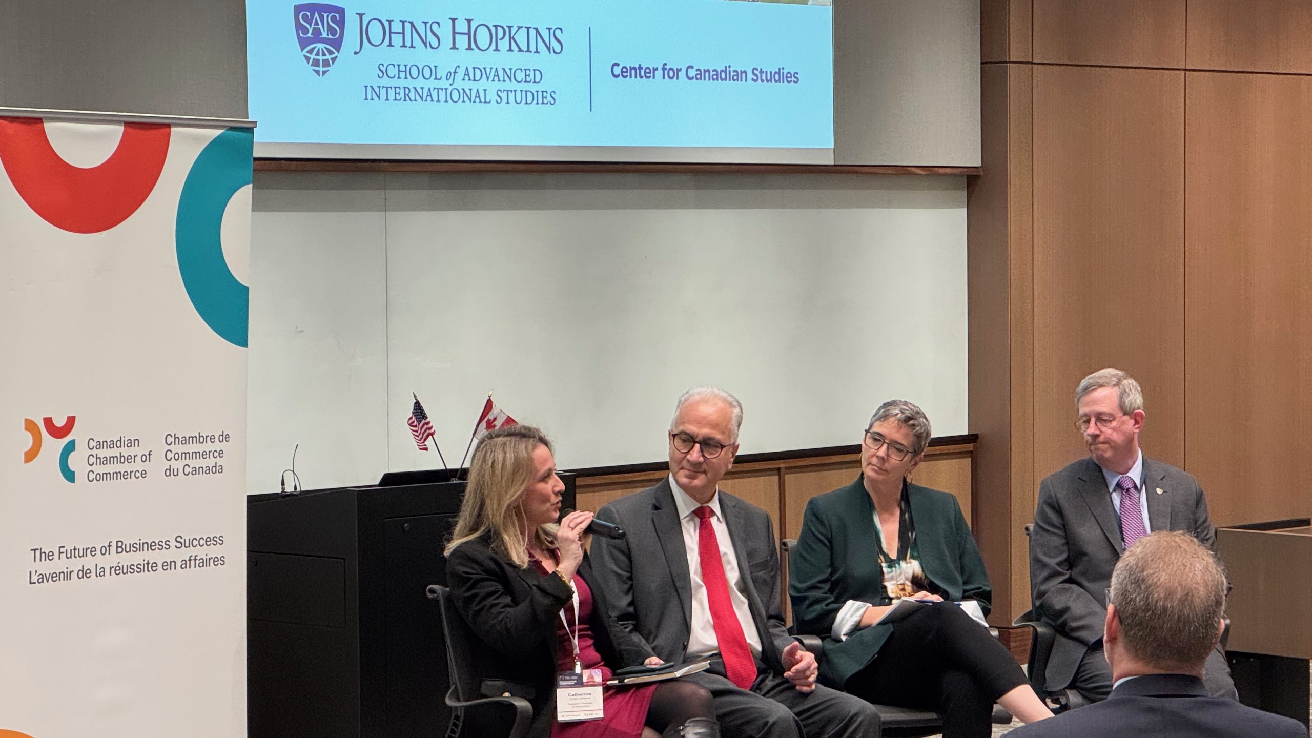 Four panelists in business attire sit and discuss on stage at a conference. Behind them are Canadian and U.S. flags, a Canadian Chamber of Commerce sign, and a Johns Hopkins Center for Canadian Studies sign.
