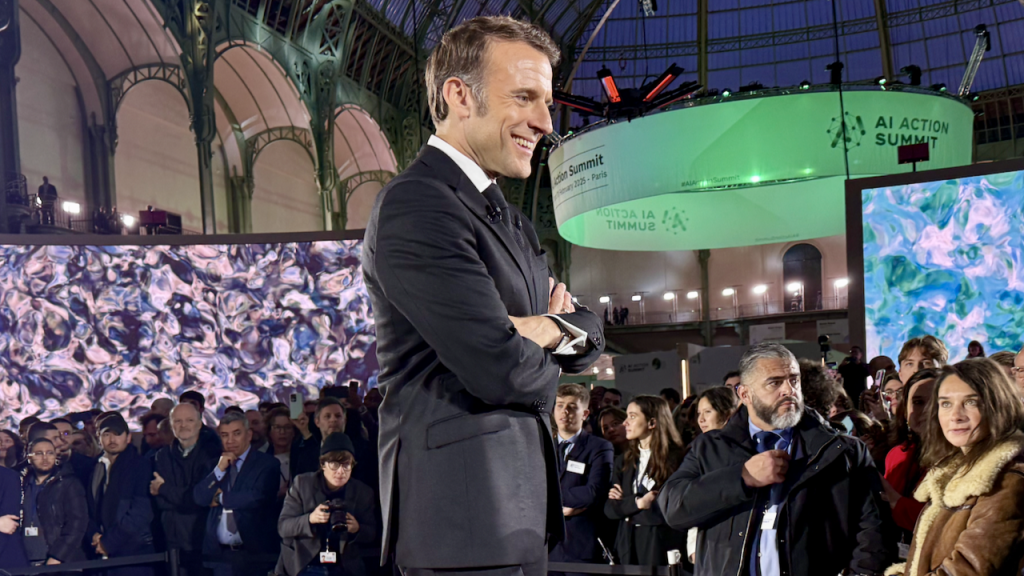 A man in a suit stands smiling with arms crossed at an indoor event with large digital screens, a crowd of people, and a green sign reading AI Action Summit in the background.