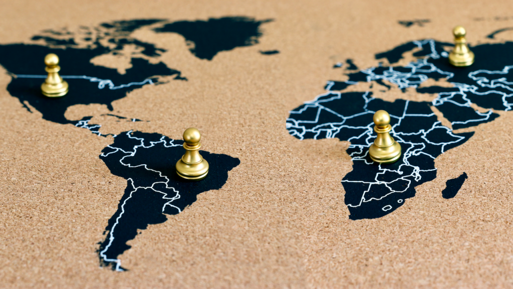 A corkboard world map with four golden chess pawns placed on North America, South America, Europe, and Africa.