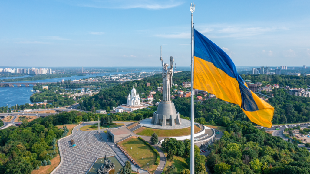 A large Ukrainian flag waves in the foreground with the Motherland Monument and surrounding cityscape of Kyiv, including trees, buildings, and a river, visible in the background under a clear blue sky.