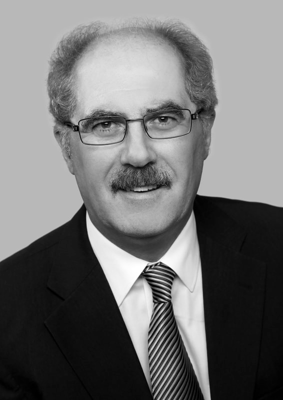A middle-aged man with gray hair, glasses, and a mustache is wearing a dark suit, a white shirt, and a striped tie, smiling slightly against a plain gray background.