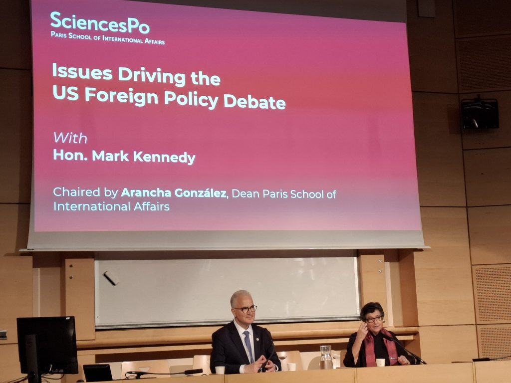 A man and a woman sit at a desk in front of a large screen displaying a SciencesPo event titled “Issues Driving the US Foreign Policy Debate” with Hon. Mark Kennedy, chaired by Arancha González at the Paris School of International Affairs.
