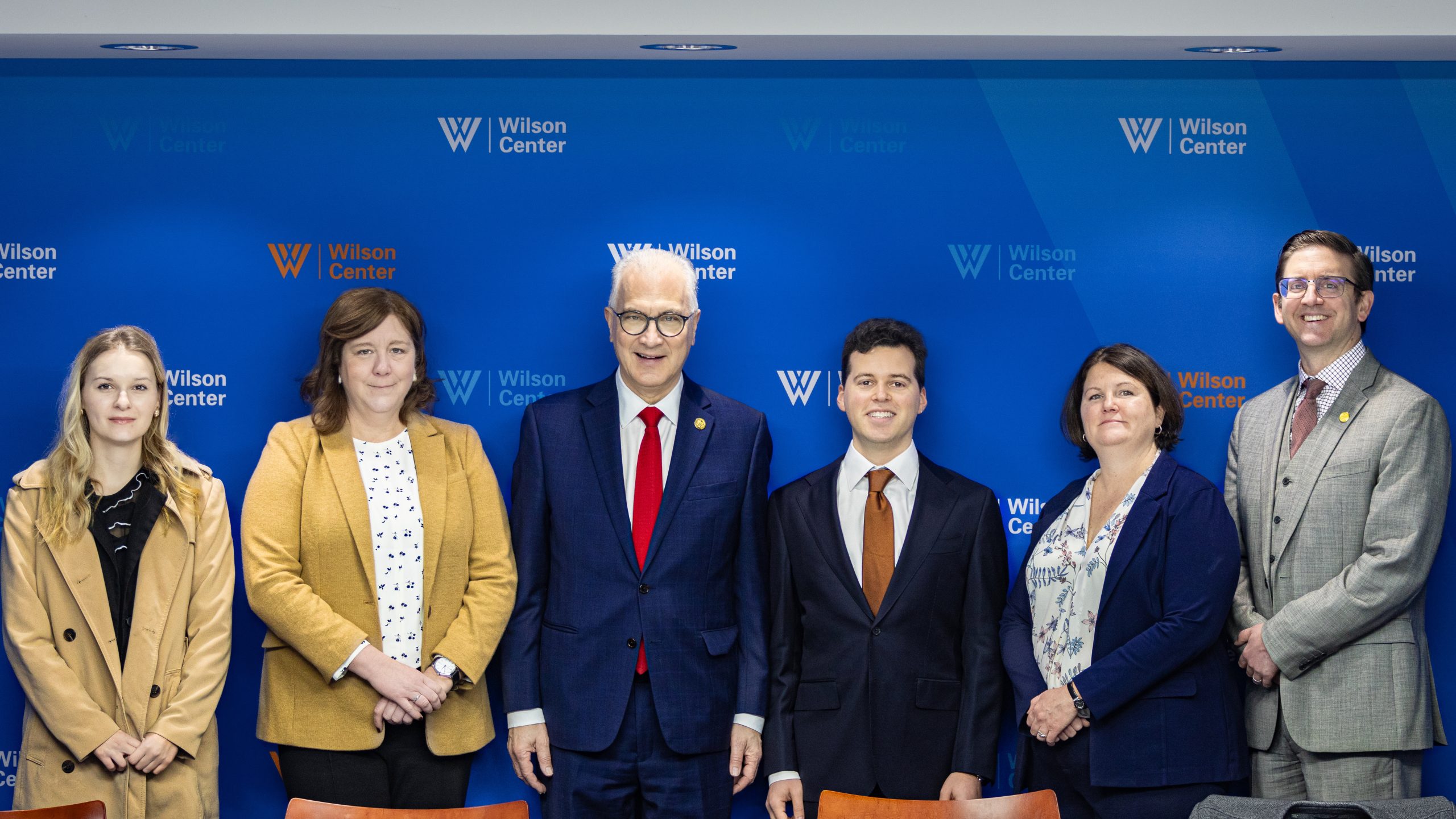 Six people in business attire stand in front of a blue backdrop with Wilson Center logos, posing for a group photo. Four men and two women are smiling, with the central man wearing a red tie and glasses.