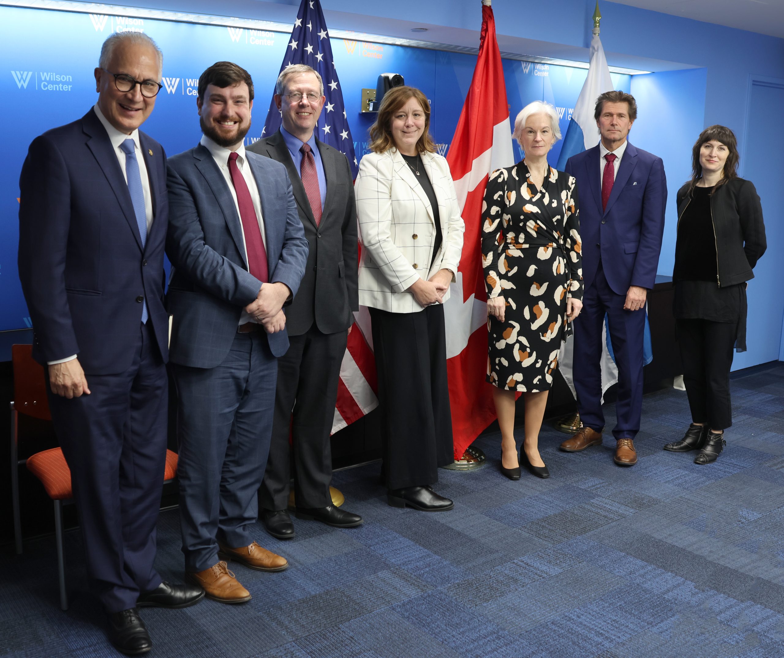 Seven professionally dressed people stand in a row, smiling, in front of American and Canadian flags and a blue wall with Wilson Center signage.