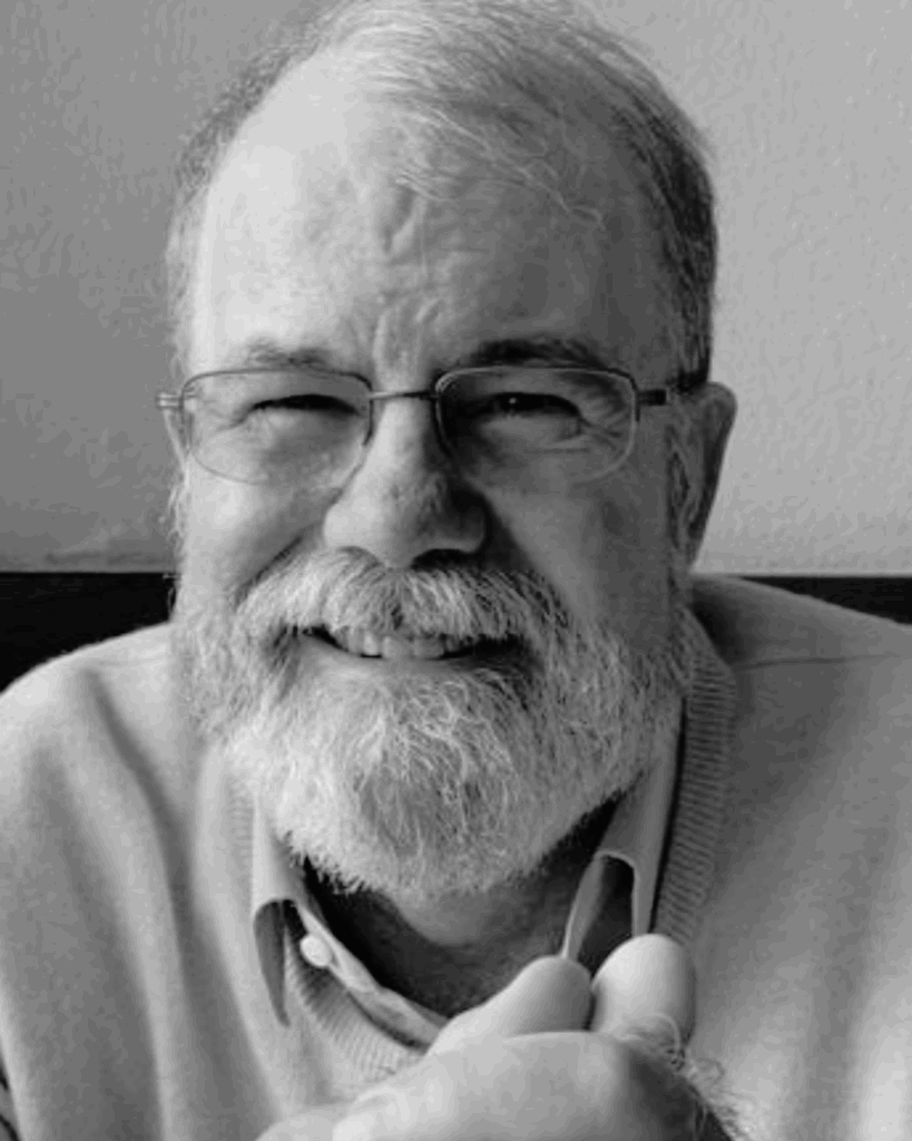 A smiling older man with glasses and a beard, wearing a light sweater, sits indoors against a plain wall; the image is in black and white.