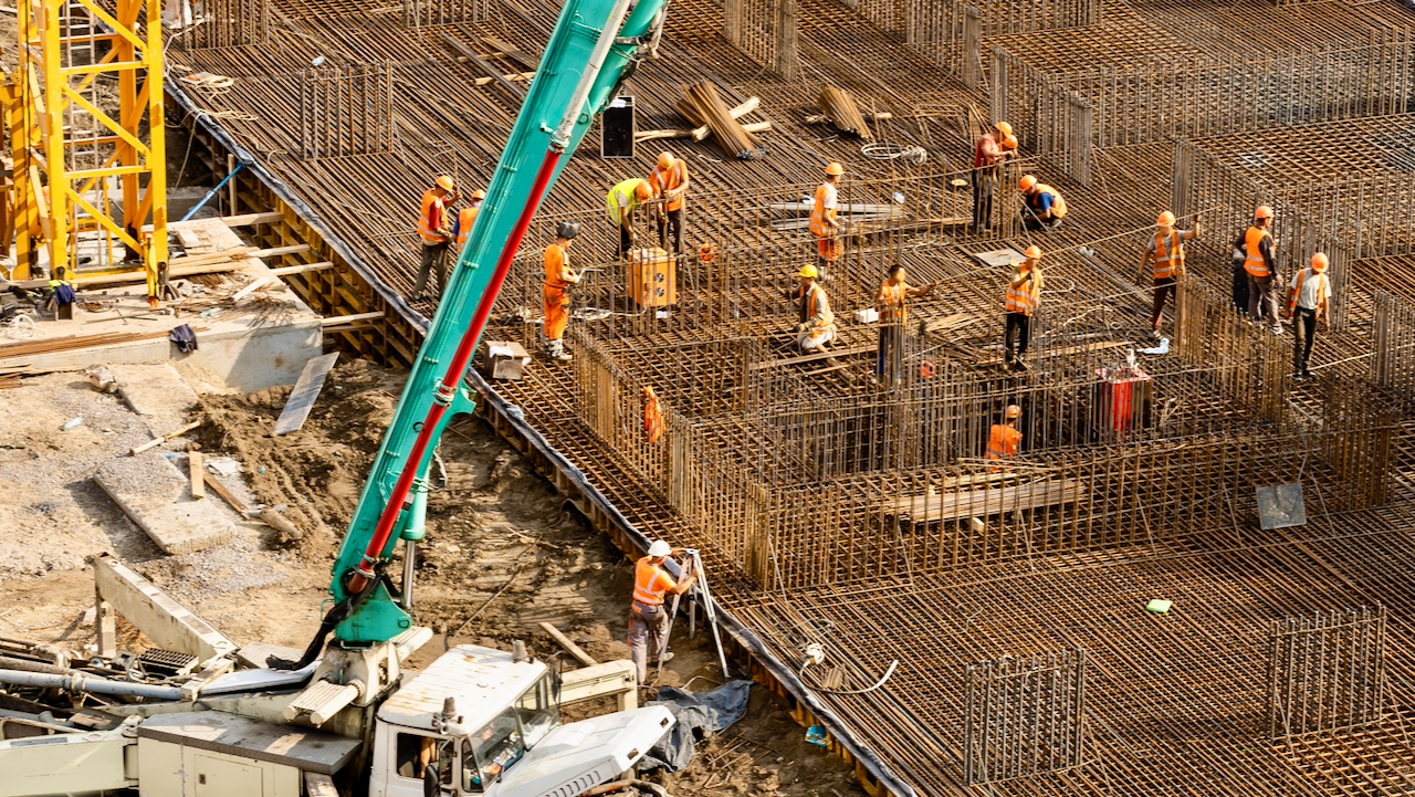Construction workers in orange safety vests and helmets are working on a large building site, assembling steel rebar frameworks with heavy machinery and equipment visible around the area.