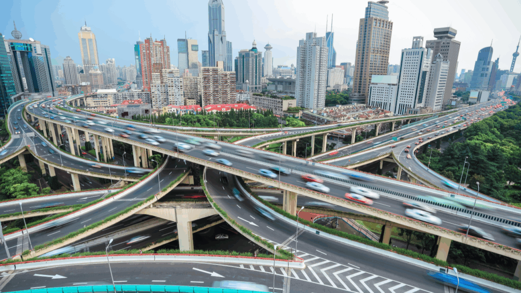 A busy multi-level highway interchange with blurred cars in motion, surrounded by green trees and modern skyscrapers in an urban cityscape under a clear sky.