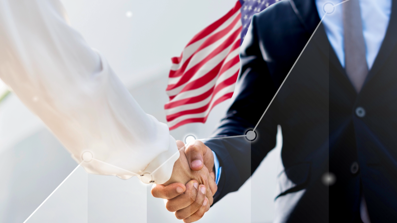 Close-up of two people shaking hands, one in a white sleeve and one in a dark suit, with a blurred American flag in the background.
