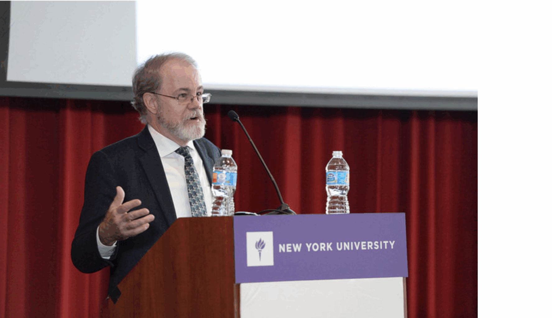A man in a suit speaks at a podium with a New York University sign, with two water bottles on the podium and a red curtain in the background. A computer screen is visible with several apps and browser tabs open.