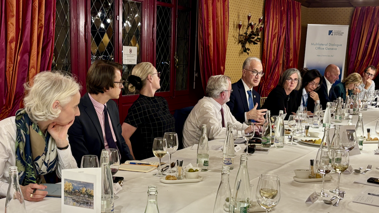 A group of people in formal attire are seated around a long dining table, engaged in discussion. The table is set with drinks, dishes, and documents, in a warmly lit room with red windows and a banner in the background.