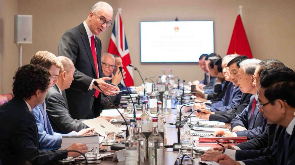 A group of officials in suits sit around a conference table during a formal meeting. Two national flags, one of the UK and one of China, are visible in the background, and the atmosphere appears serious and attentive.