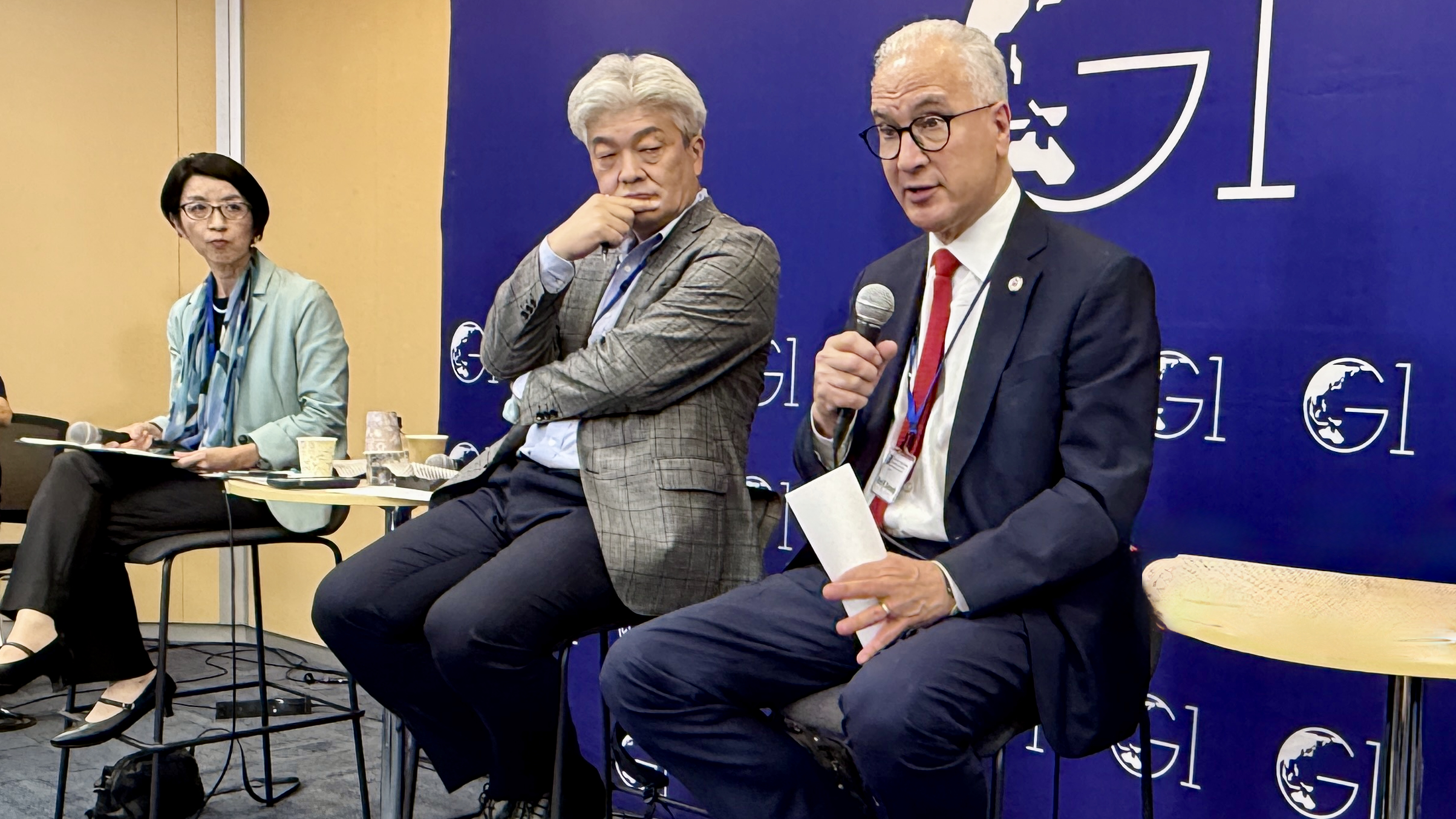 Three people sit on high chairs in front of a blue backdrop with G1 logos. One man holds a microphone and speaks, while the others listen attentively. They appear to be participating in a panel discussion or forum.