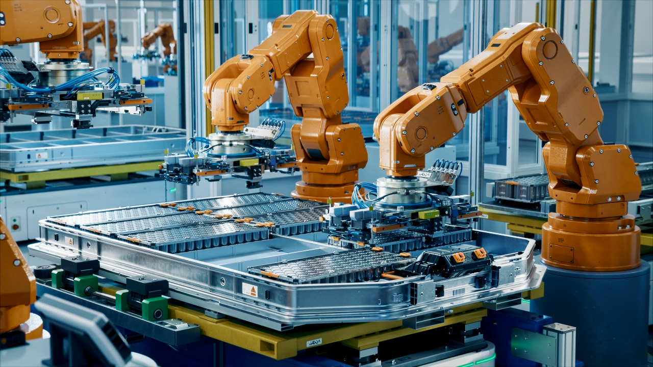 Two orange robotic arms assemble a large battery pack on an automated production line in a high-tech factory setting, surrounded by machinery and electronic components.