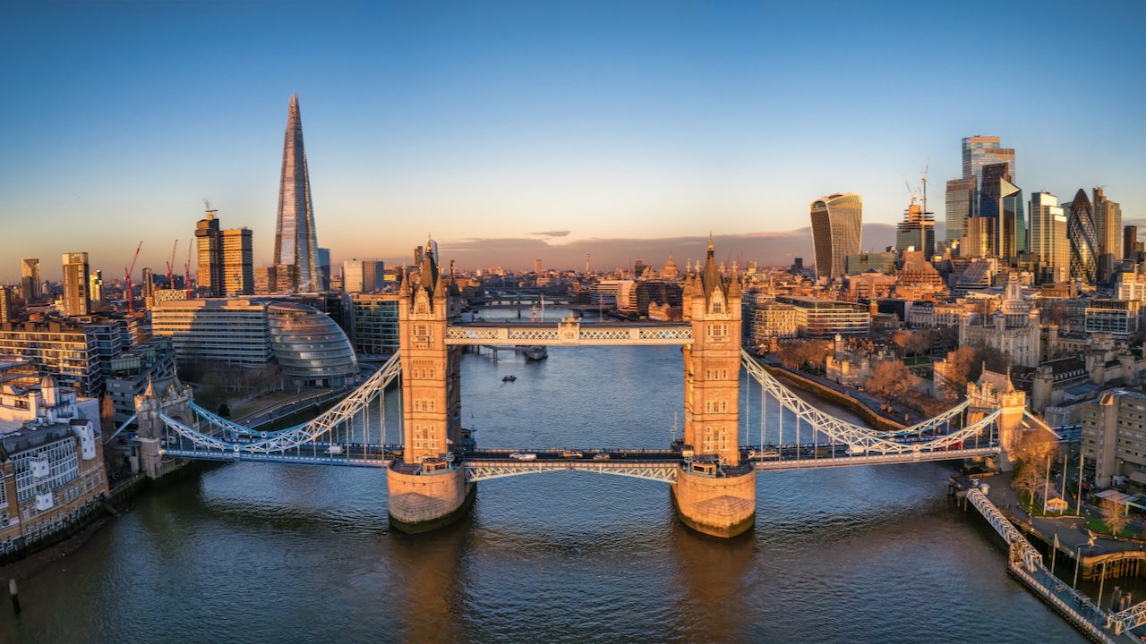 Aerial view of London’s Tower Bridge spanning the River Thames at sunset, with the Shard skyscraper on the left and modern city buildings in the background under a clear sky.