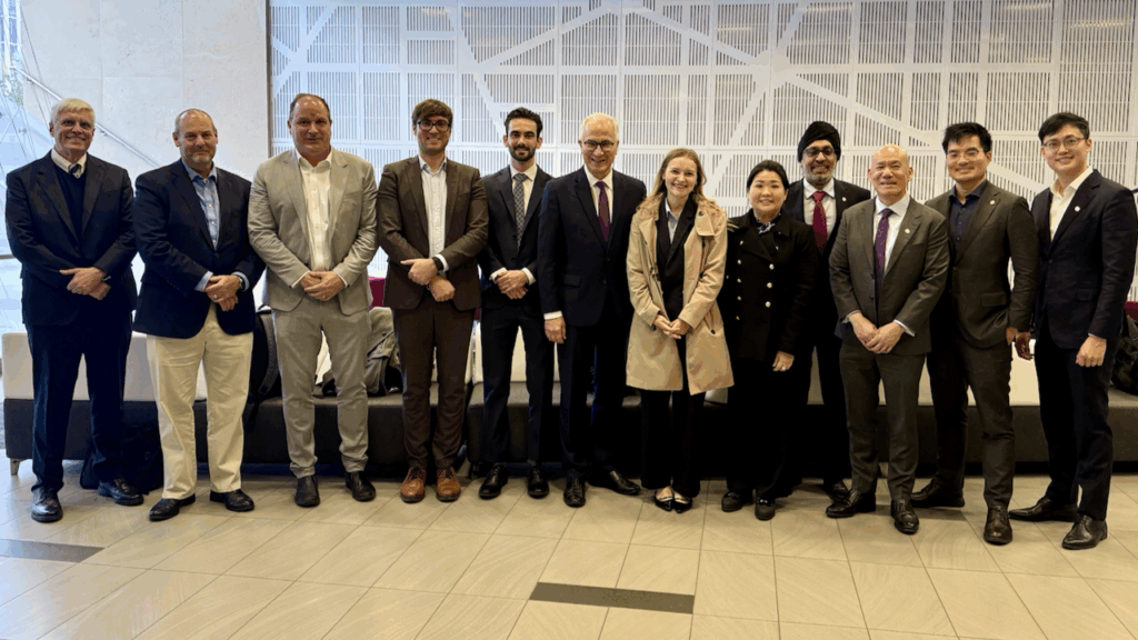 A group of twelve people, dressed in business and formal attire, stand indoors in a row, smiling at the camera. The background features a modern, geometric wall design.