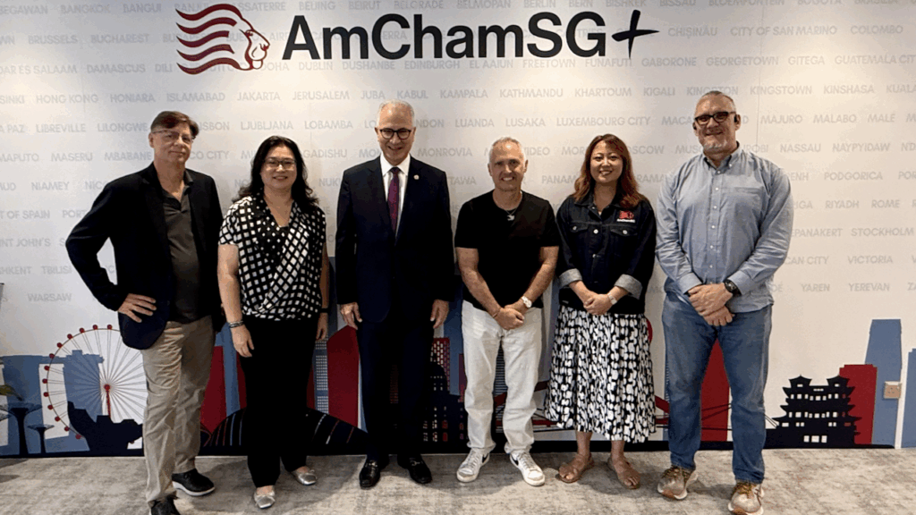 Five people stand in a row, smiling at the camera, in front of an “AmChamSG” backdrop featuring a lion logo and city names. The group includes three men and two women, all in business casual attire.