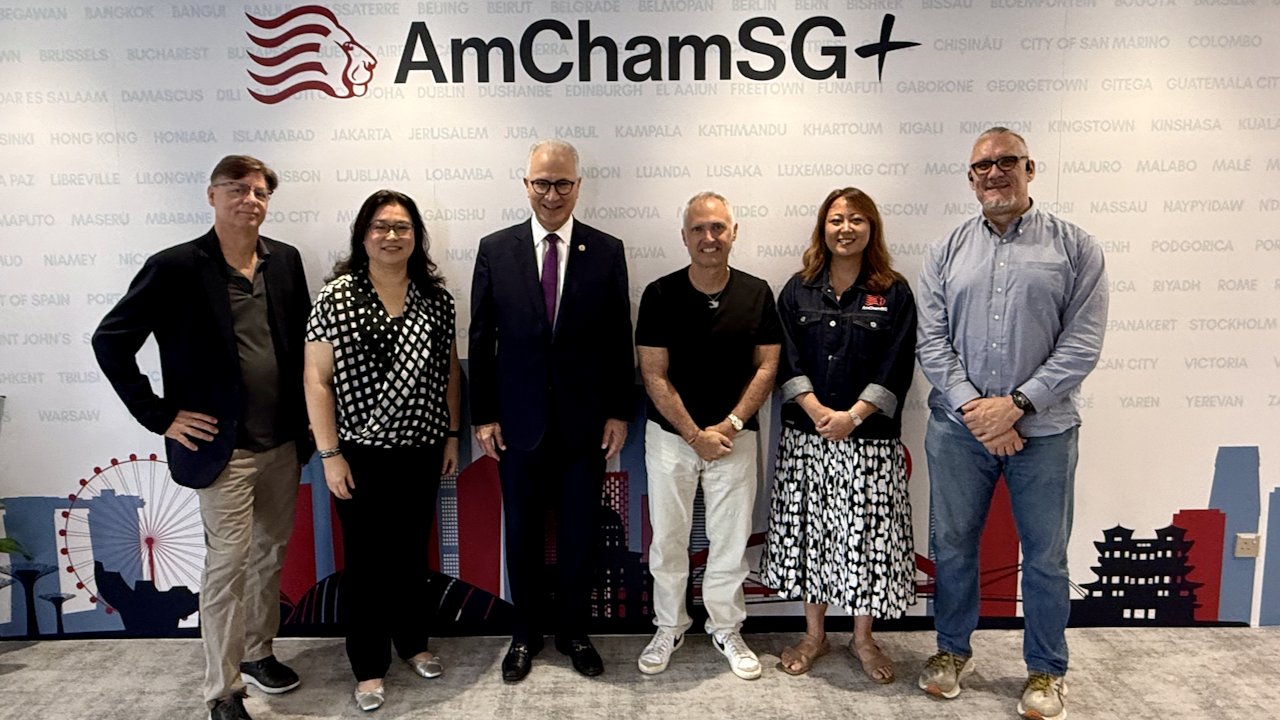 Five people stand in a row, smiling at the camera, in front of an “AmChamSG” backdrop featuring a lion logo and city names. The group includes three men and two women, all in business casual attire.
