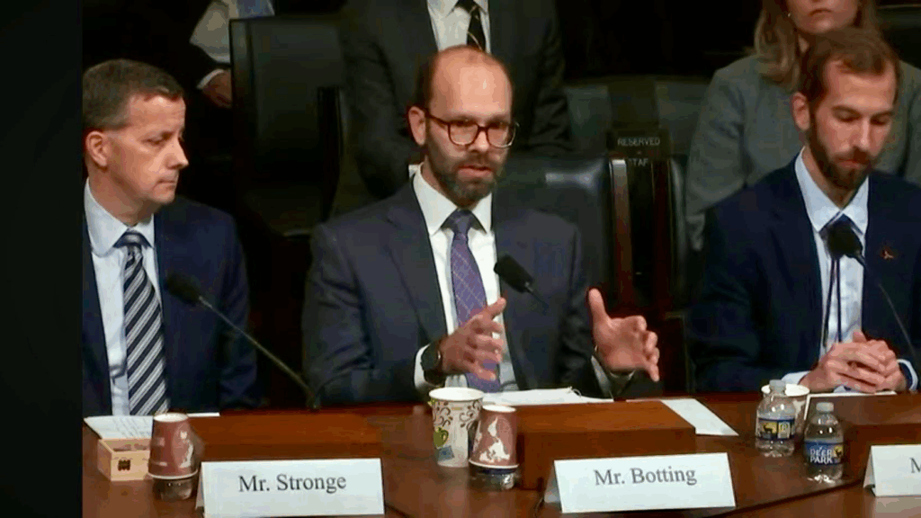 Three men in suits sit at a table during a formal meeting or hearing. The man in the center, labeled Mr. Botting, is speaking and gesturing with his hands. Nameplates and microphones are in front of each person.