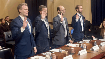 Four men in suits stand side by side with their right hands raised, appearing to take an oath at a formal hearing. They stand behind a desk with microphones, nameplates, and documents. People are seated in the background.