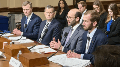 Four men in suits sit at a table with microphones, speaking and taking notes during a formal meeting or hearing. Several people sit in the background, listening attentively.