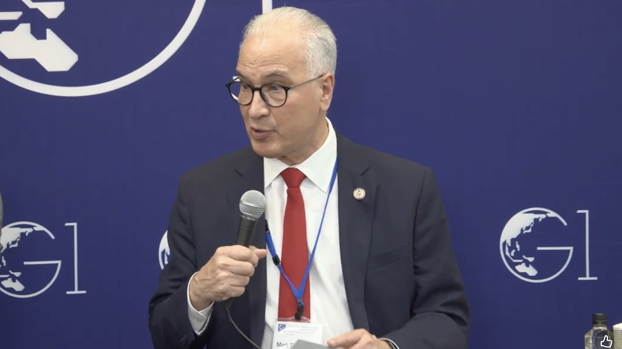 A man in a suit, red tie, and glasses speaks into a microphone at a panel event. He wears a conference badge and sits in front of a blue backdrop with globe and G1 logos.