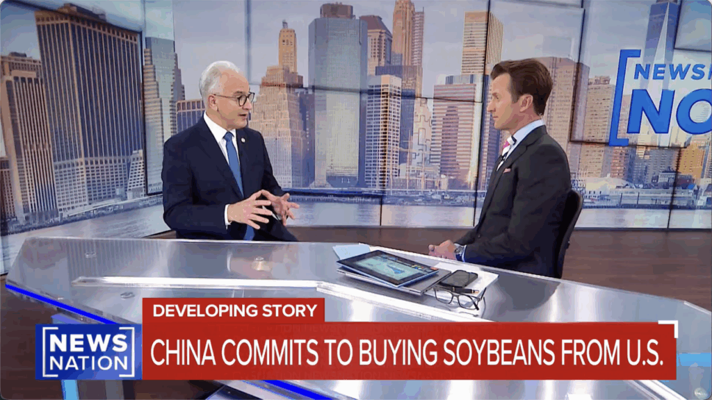 Two news anchors sit at a glass desk in a studio with a city skyline backdrop. The headline reads, CHINA COMMITS TO BUYING SOYBEANS FROM U.S. on the screen, with NEWSNATION branding visible.