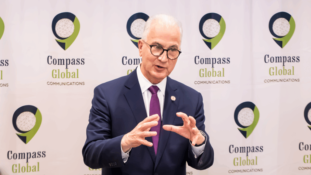 A man in a navy suit and purple tie gestures while speaking in front of a Compass Global Communications branded backdrop.
