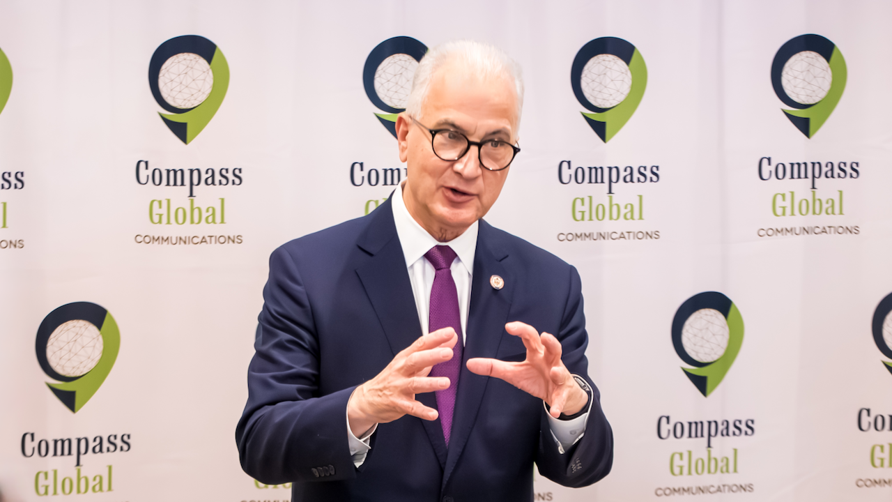 A man in a navy suit and purple tie gestures while speaking in front of a Compass Global Communications backdrop.