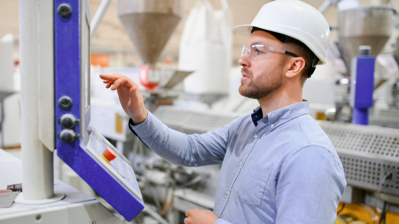A man wearing a white hard hat, safety goggles, and a light blue shirt operates a control panel in an industrial or manufacturing facility. Machinery and equipment are visible in the background.