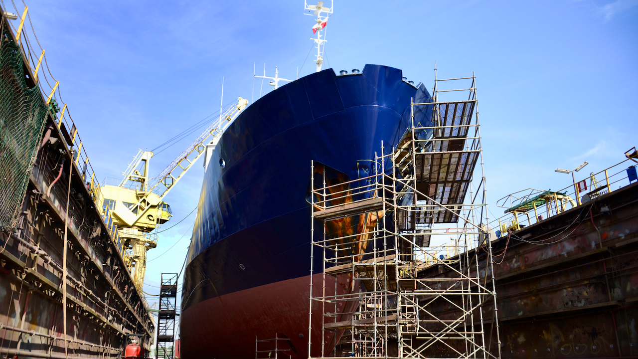 A large ship in dry dock is surrounded by scaffolding for maintenance or repairs, with a crane and equipment visible in the background under a clear blue sky.