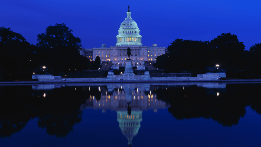 The U.S. Capitol building is illuminated at night, with its lights reflecting clearly in a calm body of water in the foreground. The sky is a deep blue, and trees frame the scene.