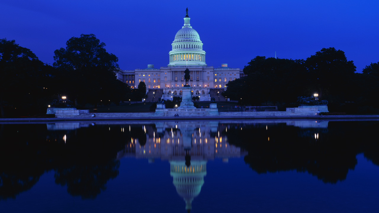 The U.S. Capitol building is illuminated at night, with its lights reflecting clearly in a calm body of water in the foreground. The sky is a deep blue, and trees frame the scene.