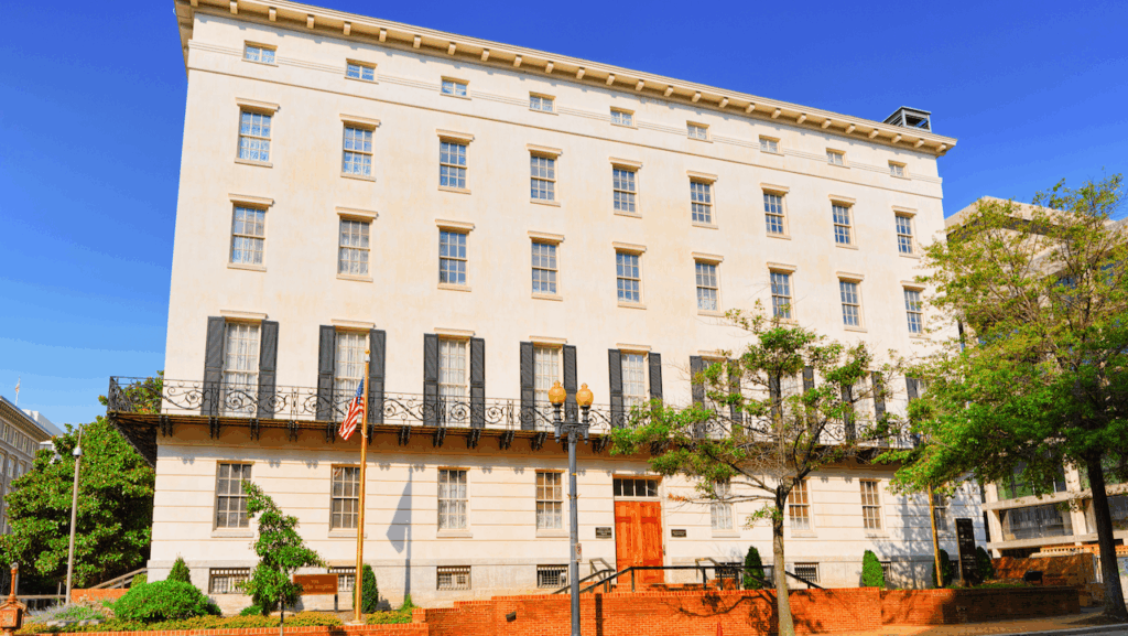 A large, cream-colored, five-story historic building with black balcony railings, tall windows, and an American flag outside, set against a clear blue sky with trees and a brick wall in the foreground.