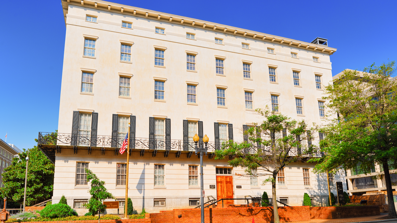 A large, cream-colored, five-story historic building with black balcony railings, tall windows, and an American flag outside, set against a clear blue sky with trees and a brick wall in the foreground.
