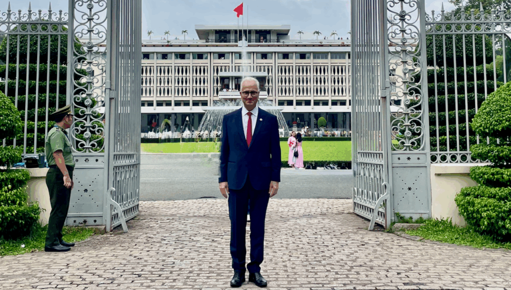 A man in a dark suit and red tie stands smiling between open iron gates, with a large modern building, green lawn, and a red flag in the background. People are visible in the distance near the building.