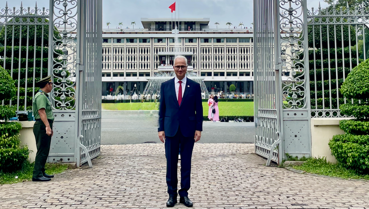 A man in a dark suit and red tie stands smiling between open iron gates, with a large modern building, green lawn, and a red flag in the background. People are visible in the distance near the building.