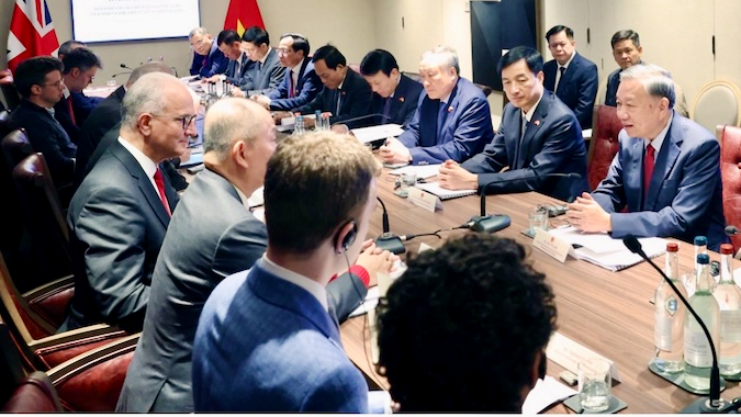 A group of officials in business attire sit around a long conference table engaged in discussion. Papers, microphones, and water bottles are on the table. Flags and a screen are visible in the background.