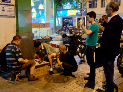Three men play an outdoor board game at night on a city sidewalk, while three onlookers, including one taking photos with a phone, watch them. Bicycles and shopfronts are visible in the background.