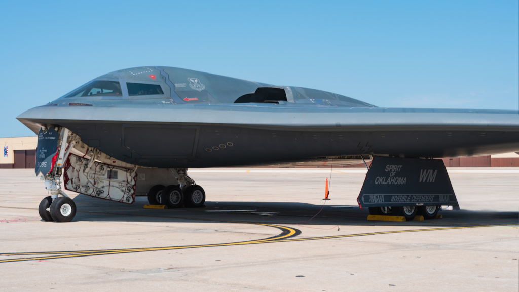 A B-2 Spirit stealth bomber, labeled Spirit of Oklahoma, parked on an airbase tarmac under a clear blue sky, with its landing gear exposed and wheel chocks in place.