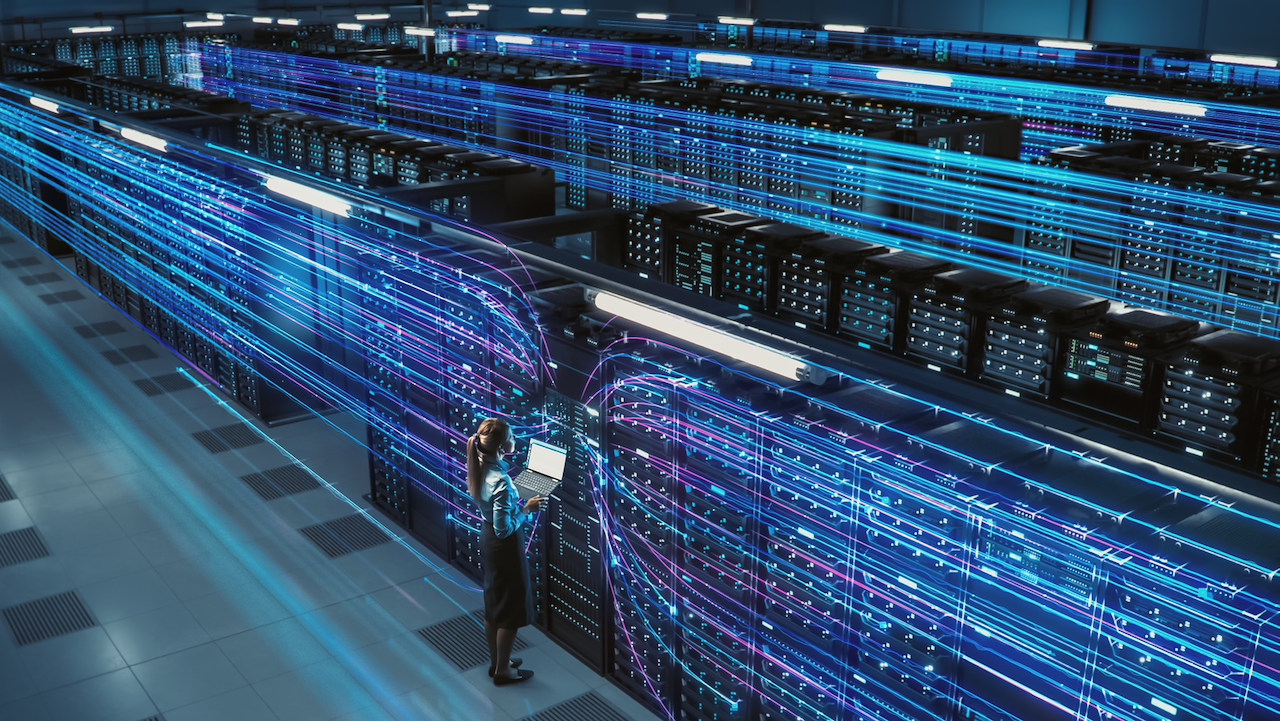 A woman stands with a laptop in a large server room, surrounded by rows of servers and blue digital light trails representing data transfer throughout the facility.