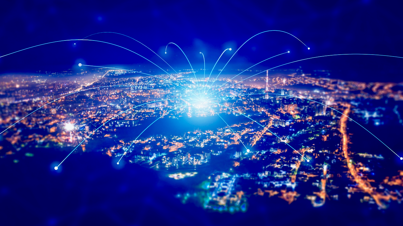A glowing cityscape at night with blue light trails and arcs connecting various points, symbolizing digital connectivity or data transfer across an urban network.