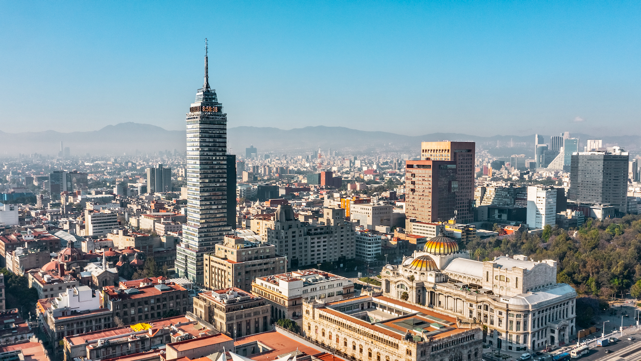 Aerial view of Mexico City’s skyline featuring the Torre Latinoamericana, the Palacio de Bellas Artes, and various modern and historic buildings under a clear blue sky with mountains in the distance.