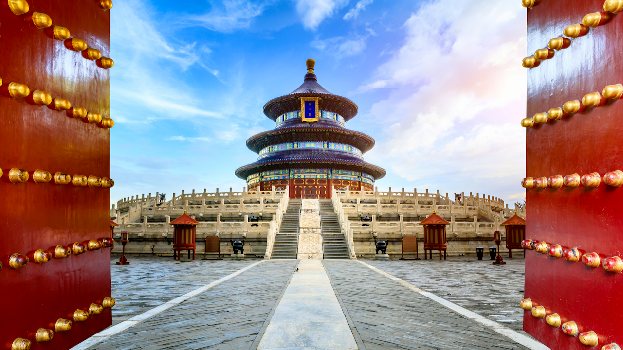 The Temple of Heaven in Beijing, China, viewed through open red doors with golden studs, under a bright blue sky with scattered clouds.